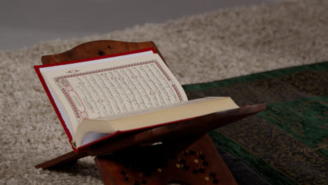 Nahaufnahme-Einer-Offenen-Kopie-Des-Korans-Auf-Einem-Ständer-Neben-Einer-Gebetsmatte-Im-Muslimischen-Zuhause